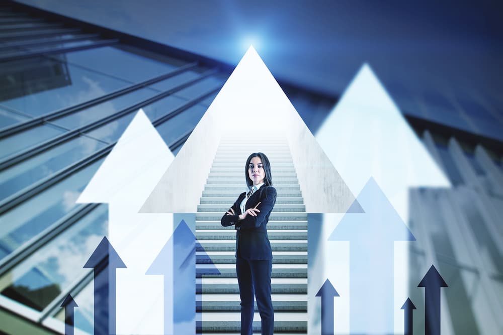 A woman stands with her arms crossed in front of a staircase and upward-pointing arrows, symbolizing career advancement.